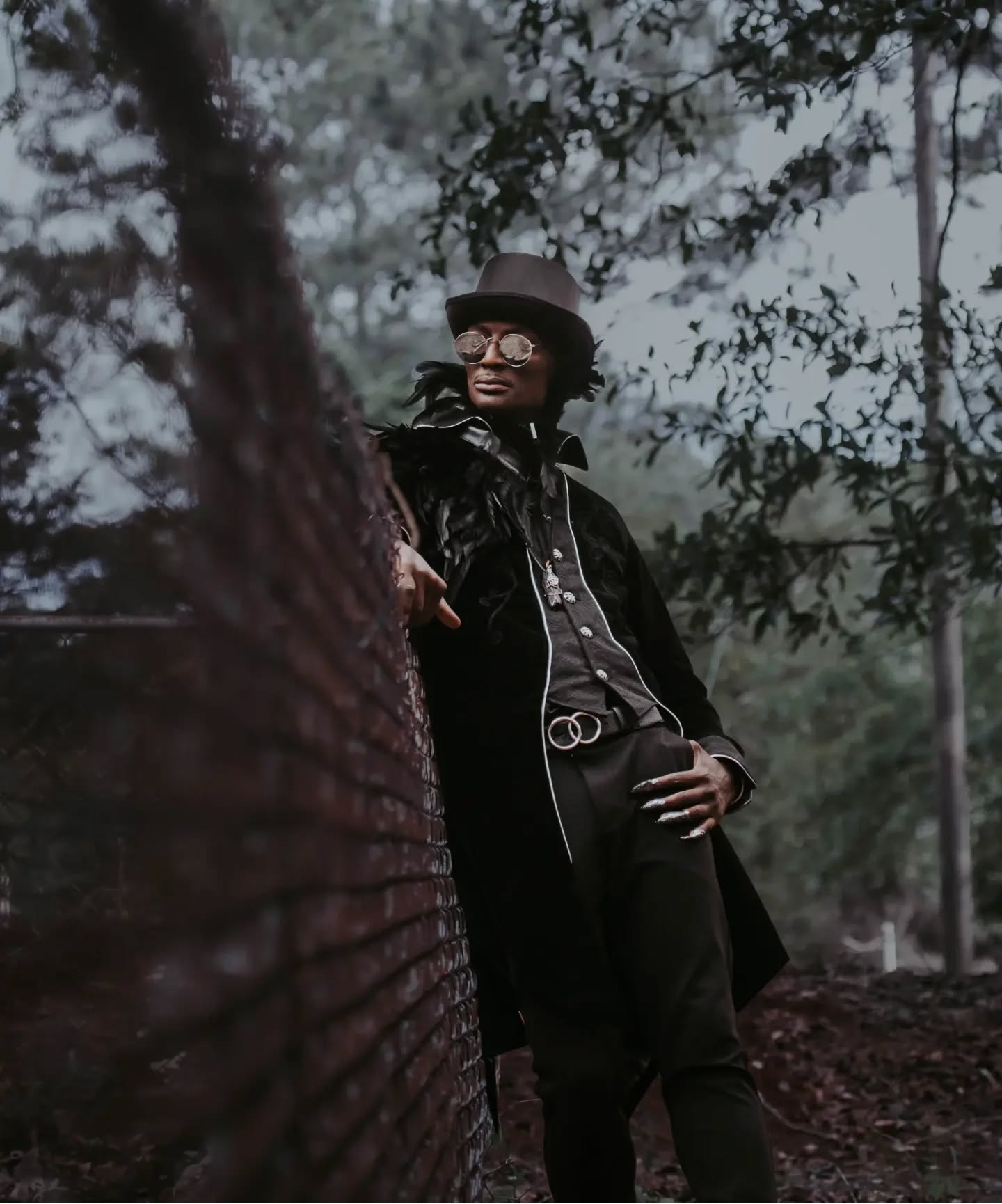 RIKKITHEFOOL in the Raven editorial wearing a top hat, feathered shoulders, round glasses and a long dark coat beside a brick column in the woods
